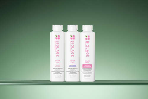 Three white bottles from the Biolage Color Last Collection, including a shampoo, a purple shampoo, and a conditioner, displayed on a clear shelf